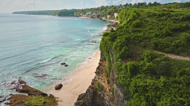 Bukit Peninsula coastline rugged cliffs, lush green vegetation and a pristine sandy beach with turquoise ocean water gently washing over shore rocks, serene tropical landscape. Aerial view flight