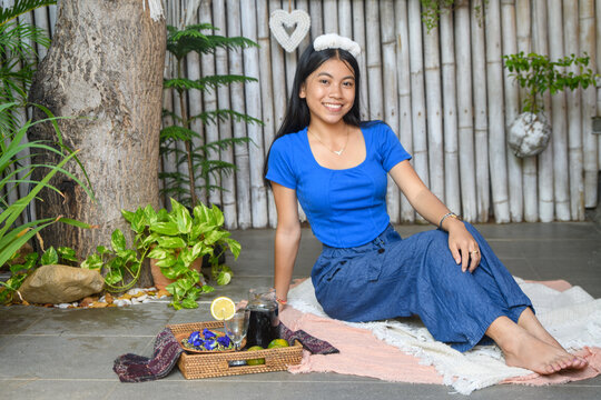 Asian woman in blue outfit relaxing on picnic blanket outdoors with butterfly pea tea tray and lemon in tropical garden