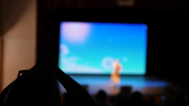 Silhouette of a woman in the audience applauding a performer on stage. Blurred view of a concert performance with a bright screen in the background