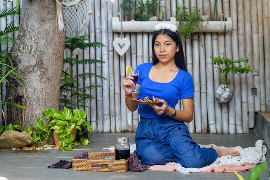 Cheerful Asian woman in blue top and jeans sitting on floor laughing while holding butterfly pea tea glass