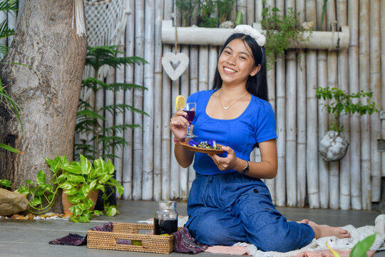 Asian woman in blue casual outfit sitting on picnic mat with butterfly pea tea and snacks in tropical bamboo garden