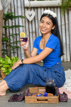 Smiling Asian woman in blue casual wear enjoying butterfly pea tea outdoors in tropical garden setting