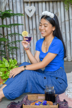 Young Asian woman in blue outfit sitting on floor holding butterfly pea tea with lemon slice and smiling