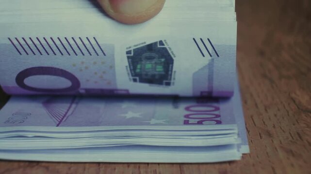 A hand is flipping through a stack of 500 Euro banknotes. The banknotes are purple and feature a security hologram.