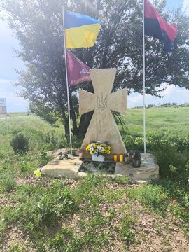 Stone cross memorial with national coat of arms trident flanked by yellow-blue, red-black and airmobile brigade flags