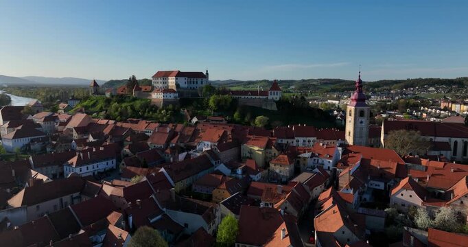 Stunning drone shot of Ptuj, Slovenia. Focuses on the dense red terracotta roofs of the old town, the historic church tower, and the majestic hilltop castle under a clear blue sky.