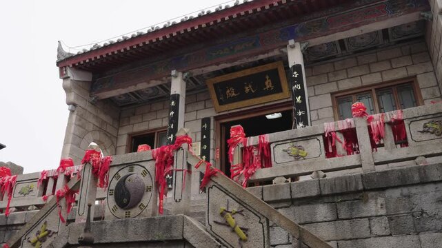 A medium close-up shot of a Taoist temple facade on Mount Hua. The stone staircase features a carved Yin-Yang bagua symbol and railings heavily adorned with red prayer ribbons.