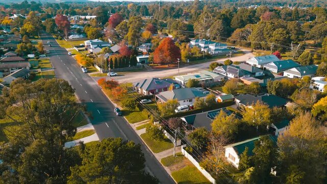 Cinematic drone footage of residential houses in Woodend, Victoria, with an upward panning motion and slight pivot revealing more of the suburban landscape, autumn trees, and moving cars on sunny morn