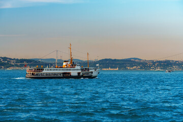 Obraz premium Traditional passenger ferry boat cruising on the blue waters of the Bosphorus strait with the cityscape and hills of Istanbul in the background during a bright sunny day.