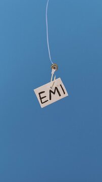 EMI word written on a paper and hooked on a fishing hook like lure. Isolated with blue background. loan repay, debt accumulation, payment crisis, liability, finance burden and pay out struggle concept