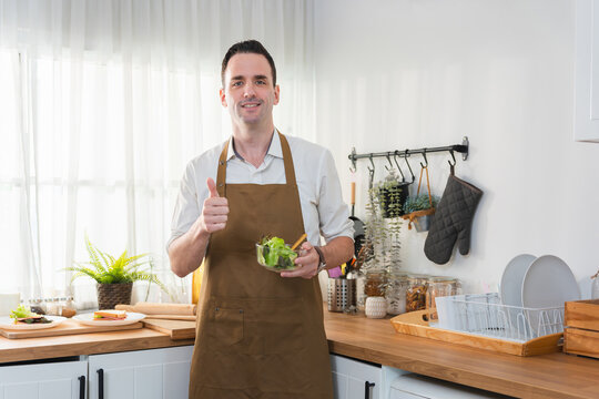 Happy man wearing apron holding a healthy salad bowl, Smiling man giving a thumbs up gesture preparing a salad at home, Cheerful man cooking healthy food in a modern domestic kitchen setting