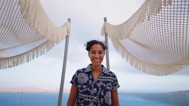 Happy young Indian woman laughing and smiling on a scenic terrace overlooking the sea in Santorini Greece. Decorative woven shade canopies frame the shot against cloudy sky and blue ocean waters.