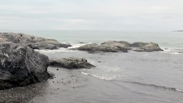 Gentle waves gracefully embrace the rugged coastline of Arctic Svalbard, Norway, on a calm, overcast day. Ancient rocks stand firm as the tide ebbs and flows, creating a serene, untamed landscape.
