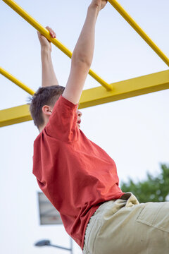 side view of a teenager boy in a red tshirt hanging and climbing on yellow metal monkey bars at an outdoor sports playground looking away