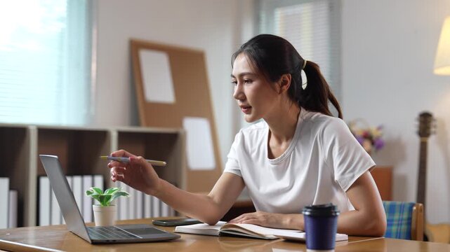 Young female student taking notes while studying online with a laptop in a cozy home environment, focused on her lesson and actively learning new concepts with a smile