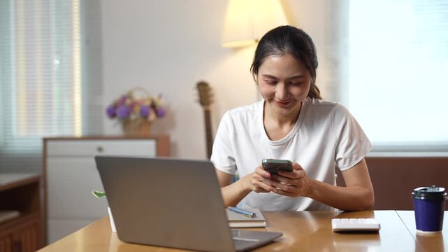 Smiling young asian woman procrastinating at home office using smartphone while working on laptop, happily texting with friends instead of focusing on her work or study