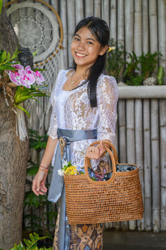 Young Balinese woman in white floral kebaya with wicker handbag against bamboo wall and dreamcatcher
