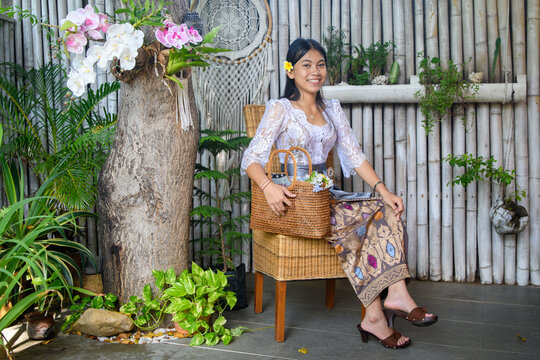 Stylish Balinese woman in floral lace kebaya sitting on rattan stool holding wicker bag in bamboo garden with orchids and dreamcatcher