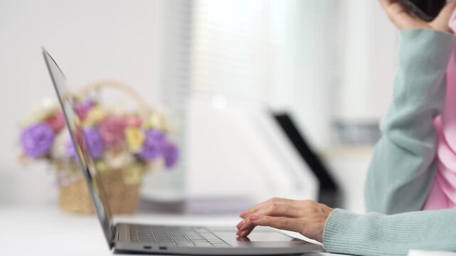 Unrecognizable young business woman scrolling with the trackpad of a laptop computer and talking on the phone in a modern office, showcasing a typical workday scene