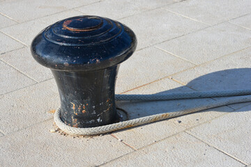 A heavy black metal mooring bollard with a secure rope wrapped around its base on a paved stone pier. © nahhan