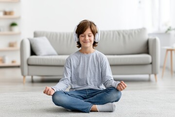 Obraz premium Young boy with headphones sits cross-legged on a carpet in a modern living room, practicing meditation with a calm expression, while a light-colored sofa and shelves are visible in the background