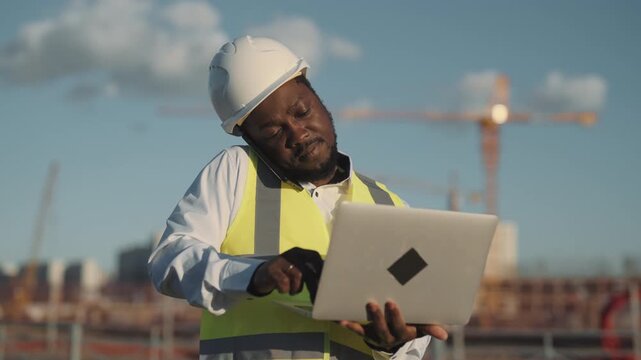 Multitasking foreman in construction site, black man work with laptop and phone. Portrait of professional architect or builder against building under construction, innovative machinery for build