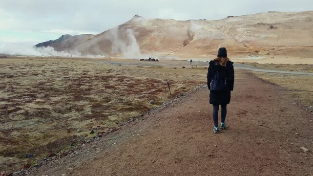 Following shot of a woman with a backpack walking along a trail in Hverir, a geothermal area in Iceland with steaming hot springs