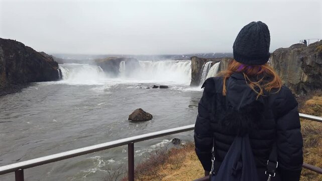 Red haired woman wearing a winter jacket and hat, admiring the powerful Godafoss waterfall in Iceland from a viewpoint on a cold day