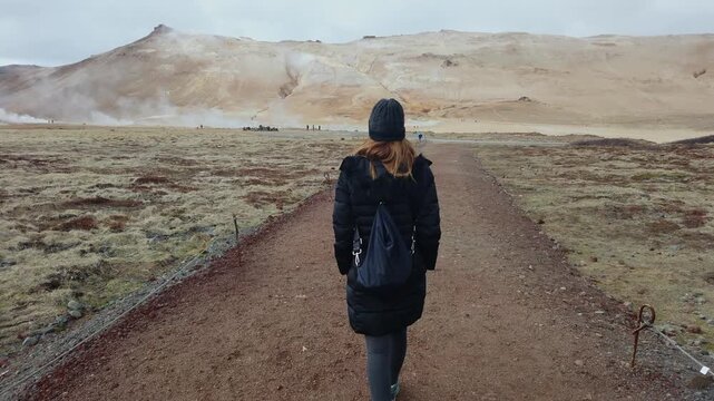 Woman traveler is trekking along a trail in the Hverir geothermal area by Lake Myvatn, Iceland, surrounded by volcanic peaks and thermal steam vents