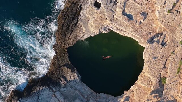 Overhead drone view of a woman swimming in the turquoise waters of Giola natural pool, surrounded by rugged rock formations on the coast of Thassos.