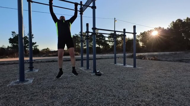 Front view of an athletic man doing pull-ups on a bar in an outdoor fitness area during sunrise. Natural backlight with trees in the background. Concept of bodyweight training and morning routine