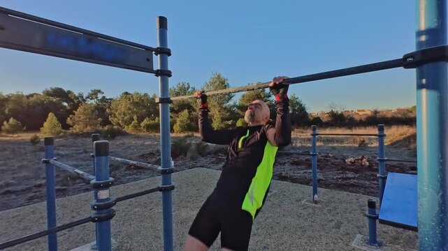 Close-up, three-quarter view of an athletic man performing pull-ups on a bar in an outdoor workout area. Warm morning light with trees in the background. Concept of discipline and physical strength.