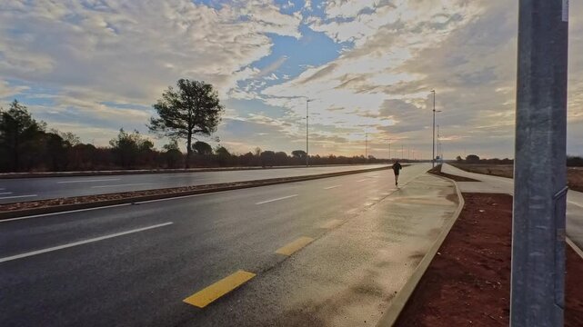 Wide shot of an athlete jogging away on a wet asphalt road toward the horizon. Dramatic morning sky and reflections. Concept of endurance, freedom, and personal journey.