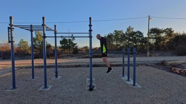Side view of an athletic man doing triceps dips on parallel bars in an outdoor fitness area. Morning sunlight with trees in the background. Concept of bodyweight training, strength, outdoors.