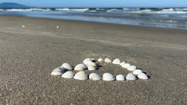 Video of a heart shape made from seashells near the sea. Romantic beach composition representing love, summer vacation, travel and coastal lifestyle.