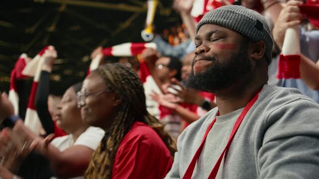 Black football supporter with drum closing eyes and holding breath in stadium stands during tense live soccer match moment.