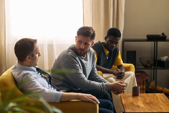 Multiethnic young adult and middle aged men sitting together, talking and listening during support meeting, expressing trust and male friendship. For counseling, community, mental health campaigns
