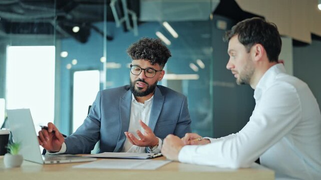 Two diverse businessmen looking at laptop screen during team discussion in office. Caucasian partner gesturing and explaining project details to surprised or focused Middle Eastern coworker.