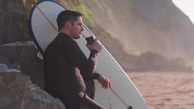 Surfer wearing a wetsuit relaxing while drinking mate on the beach at sunset