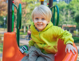 Cheerful toddler boy smiling while playing on a colorful playground slide in a bustling urban park in fall © Evgenii Bakhchev