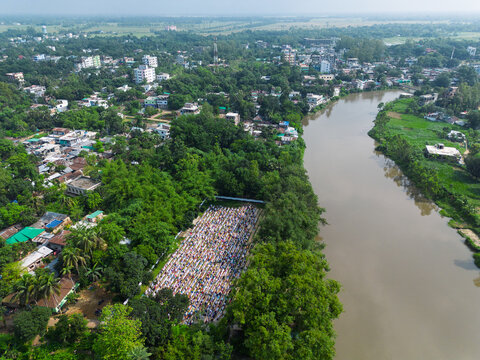 Joypurhat, Bangladesh - 28 May 2026: Aerial view of a large crowd of people gathered for prayer in an open field surrounded by dense green trees and a winding river.