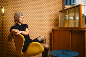 Obraz premium Woman sitting on yellow chair in vintage room with wooden furniture and decorative wall