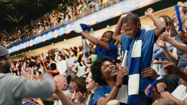 Diverse blue team football supporters chanting in packed stadium stands during live soccer match, fan scarves and excited crowd atmosphere, father, mother and son sharing family fun with laughter.