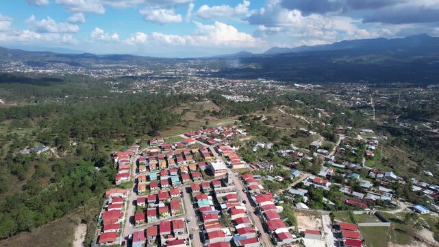 A high-altitude drone shot reveals a Latin American residential development with approximately 150 homes featuring red zinc roofs, nestled between dense green forest and open land.