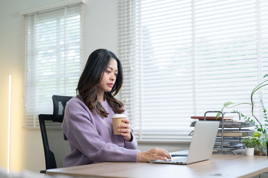 Tired asian woman holding coffee cup looking at laptop while sitting at wooden table in home office