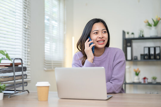Happy asian woman talking on phone call over a laptop and coffee as sitting at wooden table in home.