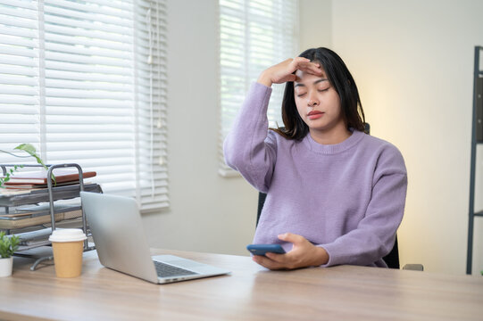 Asian woman holding phone massaging temple beside laptop as sitting at wooden table in home office.
