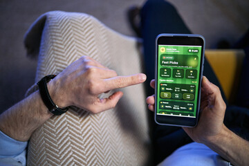 Close up of young adult man holding smartphone and tapping sports betting app, checking live odds and match picks. Useful for online gambling, mobile gaming, fintech marketing © DragonImages