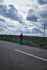 Obraz premium Adult man standing in a rural agricultural field observing wind turbines under a dramatic cloud-filled sky. Concept of renewable energy, sustainability, clean power generation, energy transition