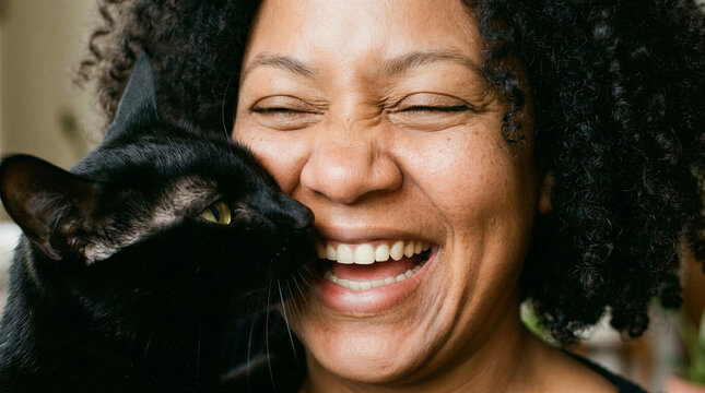 A dark-skinned, full-figured woman smiles while hugging her black cat indoors.  